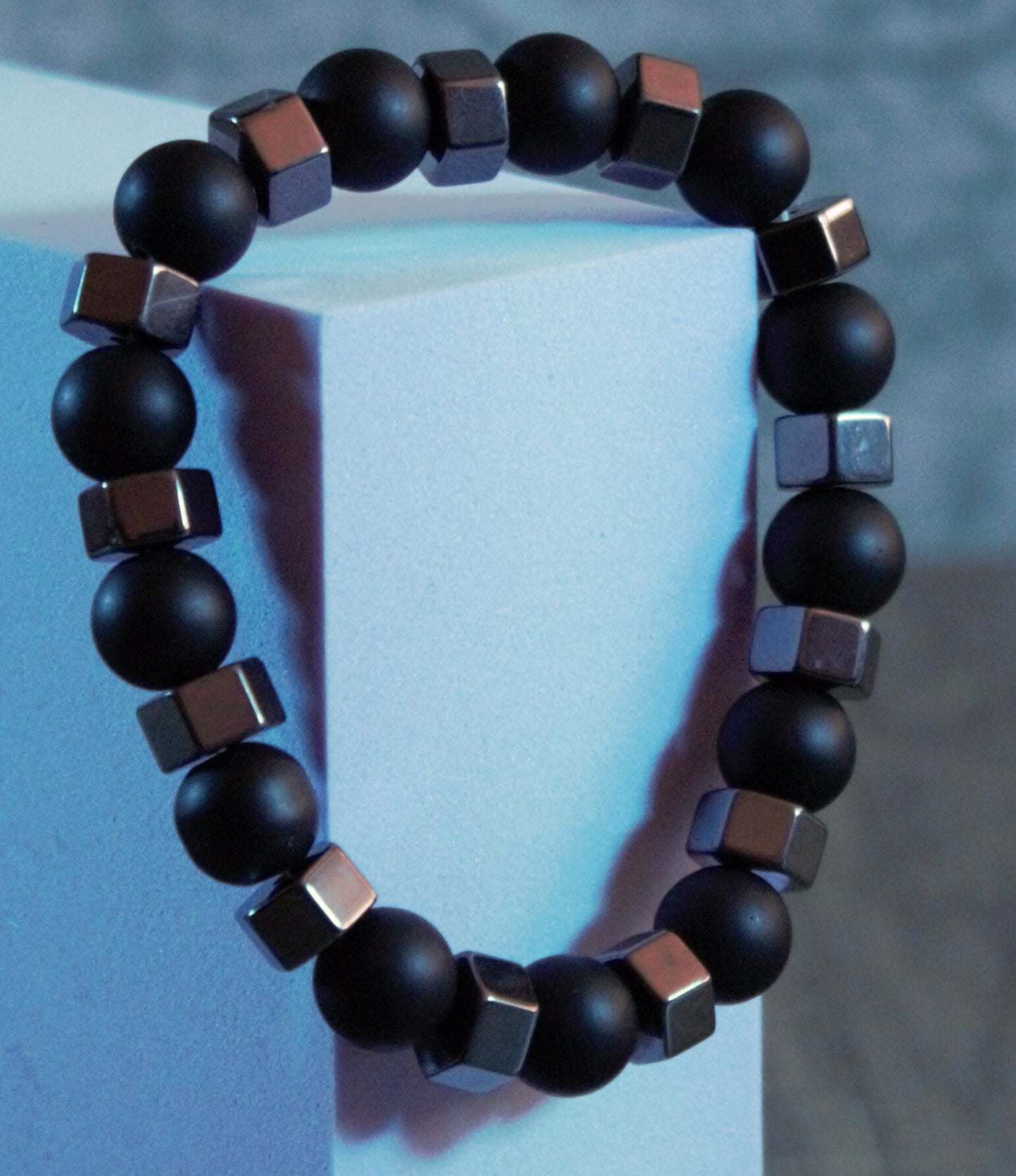 Matte black beaded bracelet with metallic cube accents, styled on a geometric pedestal under ambient moody lighting – Midnight Pulse by Charmed Curations.